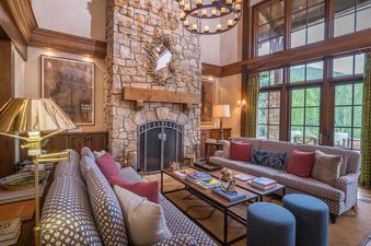 A spacious living room with a stone fireplace, large windows overlooking a forested area, and comfortable furniture.
