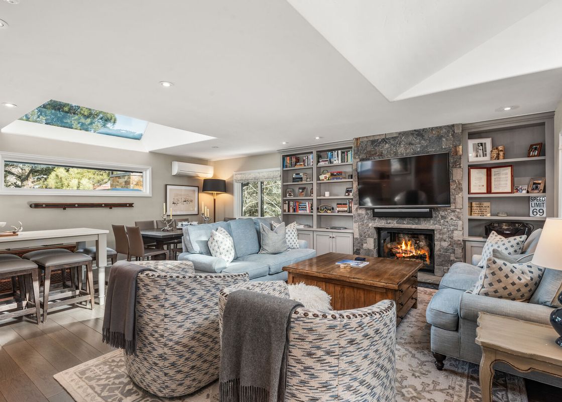A bright and cozy living area with light blue sofas, patterned armchairs, a stone fireplace, and a large skylight.
