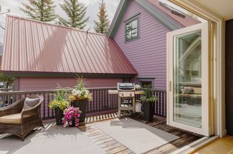The Smugglers Hideaway deck features a BBQ grill, a wicker chair, and potted flowers, set against the backdrop of the house's purple siding and a red-brown metal roof.