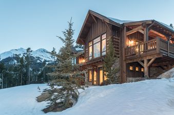 The Wolf Pack Hideaway, a rustic timber and stone home, is illuminated from within on a snowy evening with mountains and pine trees in the background.