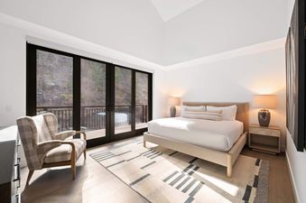 A serene bedroom at Frontgate 429 featuring a large bed, nightstands with lamps, a plaid armchair, and glass doors leading to a balcony with mountain views.