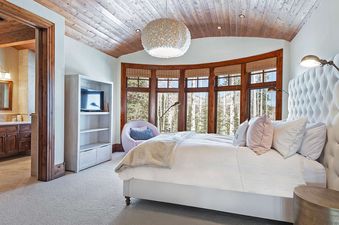 A spacious bedroom at Alpenglow at the Colony features a large white bed, a prominent bay window with forest views, and a doorway to an en-suite bathroom.