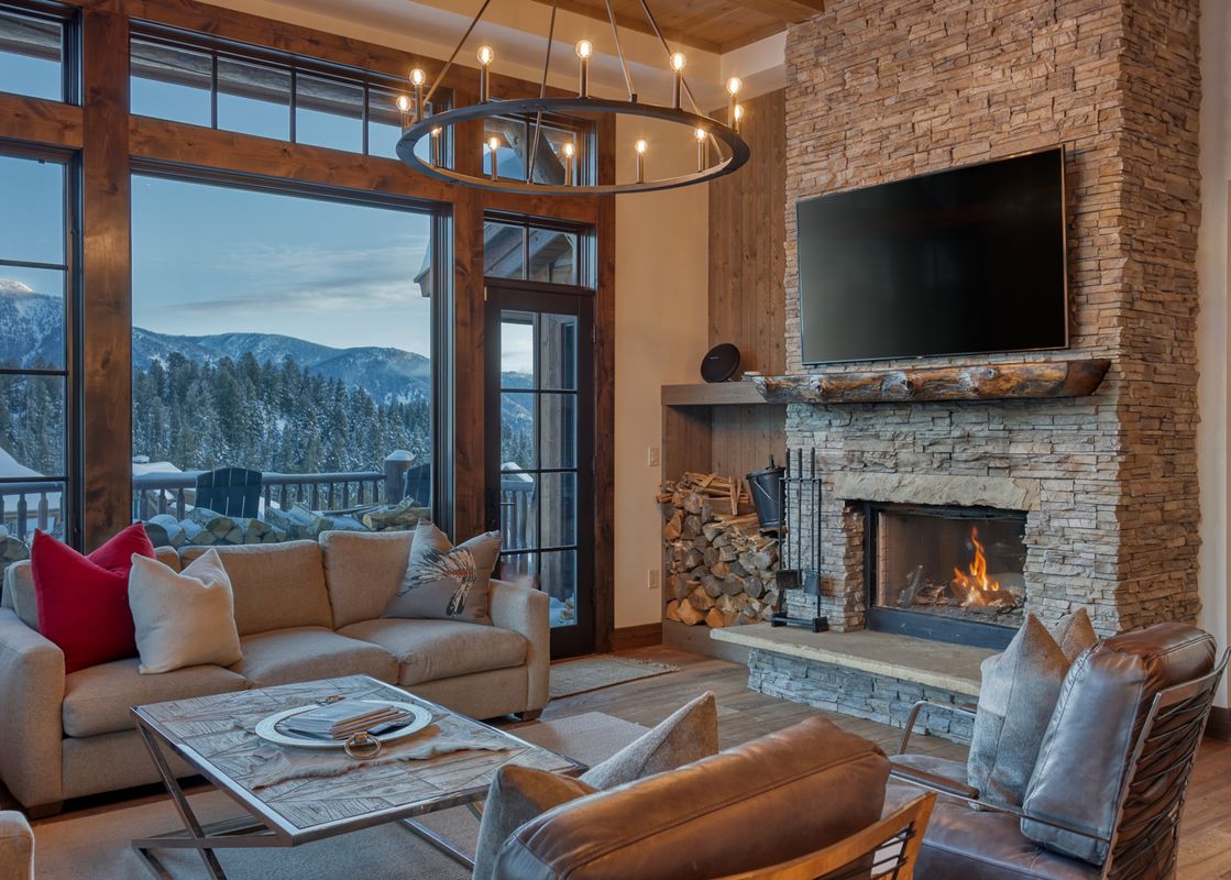 A luxurious living room in Tanager Cabin 43 features a large stone fireplace with a TV, comfortable sofas, and expansive windows offering stunning views of snow-covered mountains.