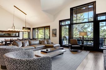 A luxurious and spacious living area in the Hummingbird Platinum 3 BR Plus Loft, featuring a large gray sectional sofa, two patterned armchairs, and floor-to-ceiling windows with views of greenery and an outdoor patio.