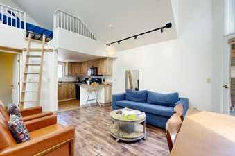 A cozy living area with a blue sofa, wooden coffee table, and a large window. The room has a loft bedroom above it.
