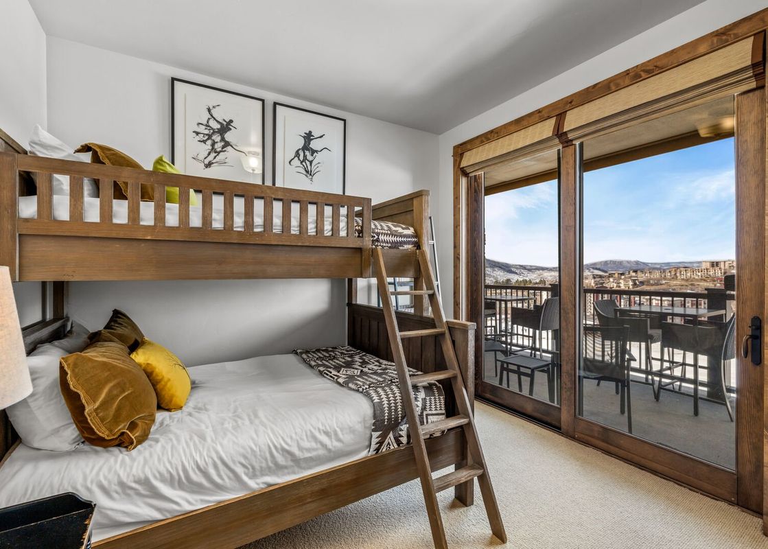 A cozy bunk room at Edgemont 2401 featuring a wooden bunk bed with white bedding, vibrant pillows, abstract art, and a balcony with an expansive view of mountains and distant buildings.