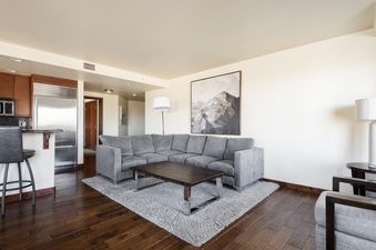 A modern open concept living space in Lowell 201B with a gray sectional sofa, a dark wood coffee table, a gray rug, and a glimpse of the kitchen with stainless steel appliances.