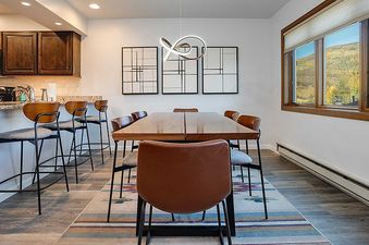 An inverted view captures the modern dining area and kitchenette of Ranch at Steamboat RA605, featuring a wooden dining table with leather chairs, abstract art, and a large window overlooking a scenic autumn landscape.