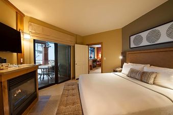 A luxurious bedroom at The Grand Summit Lodge Grand 1 King Bed features a king-sized bed, a fireplace, and a private balcony overlooking a snowy landscape.