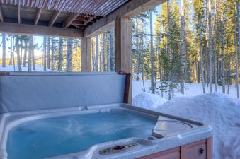 A steamy outdoor hot tub sits beneath a rustic wooden structure, surrounded by snow and a dense forest of pine trees, bathed in sunlight.