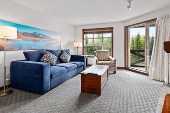 A cozy living room with a blue sofa, a wooden coffee table, and a large window offering a view of lush green trees.
