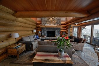 A cozy living room in The Totem Chalet with a large stone fireplace, plush sofas, and expansive windows offering a scenic view of a snow-covered forest.