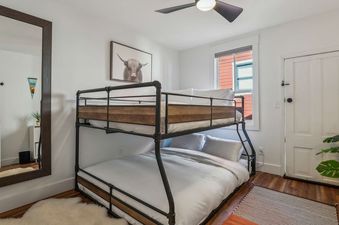 A cozy bedroom featuring an industrial-style bunk bed with white linens, a large mirror, and a window with natural light.