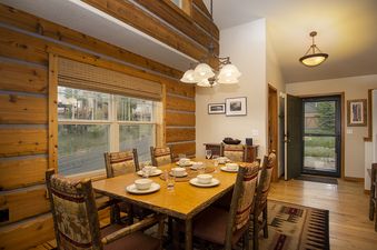 A rustic dining room features a large wooden table set for six with white dinnerware, surrounded by chairs with patterned upholstery. Exposed wooden beams adorn the walls and ceiling, while a large window offers a view of lush greenery outside.