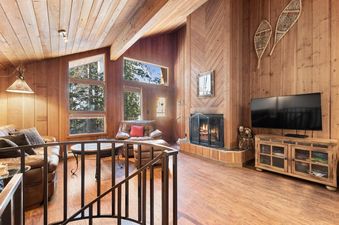 A rustic living room with wooden walls and ceiling, featuring a cozy fireplace, leather sofas, a television, and large windows looking out to trees.