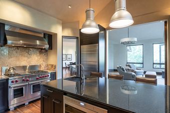 A modern kitchen with dark cabinetry, black granite countertops, and stainless steel appliances flows into a bright living room.