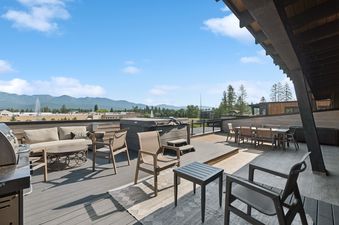 A spacious rooftop deck featuring outdoor furniture, a hot tub, a BBQ grill, and panoramic mountain views under a clear blue sky.