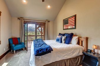 A bedroom with a view of the mountains, featuring a large bed with blue and white bedding, an armchair near the window, and an American flag hanging on the wall.