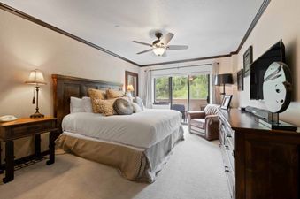 The master bedroom at St James Place 437 features a large bed with a dark wood headboard, bedside tables with lamps, a dresser with a TV, and a balcony overlooking green trees.