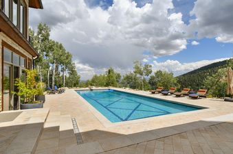 A luxury outdoor swimming pool with lounge chairs overlooking a mountain landscape under a partly cloudy sky.