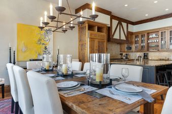 A rustic dining table set with white chairs and a chandelier, with a kitchen in the background.