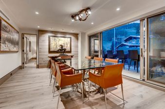 A modern dining room in Gondola One Chalet with a glass table, orange chairs, artistic lighting, and a balcony with mountain views.