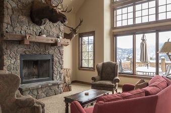 A cozy living room with a large stone fireplace, plush seating, and expansive windows offering a view of snowy mountains.