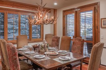 A dining room table with a wooden table, chairs, and a chandelier made from antlers. The room has large windows with wooden blinds.