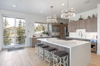 A modern kitchen with a large island, four bar stools, crystal chandeliers, stainless steel appliances, and a glass door leading to a balcony with a view of pine trees.