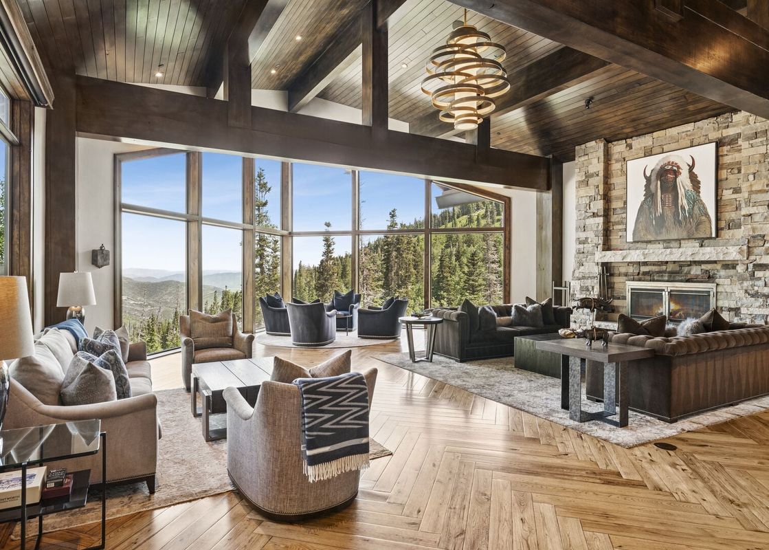 Spacious great room at Grandview at The Colony with high wood-beamed ceilings, a large stone fireplace, comfortable seating, and massive windows offering mountain views.