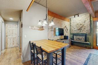 The dining area at Storm Meadows Club A216 features a rustic wooden table with four chairs, illuminated by a chandelier, next to a cozy lounge area with a stone fireplace and a wall-mounted TV.