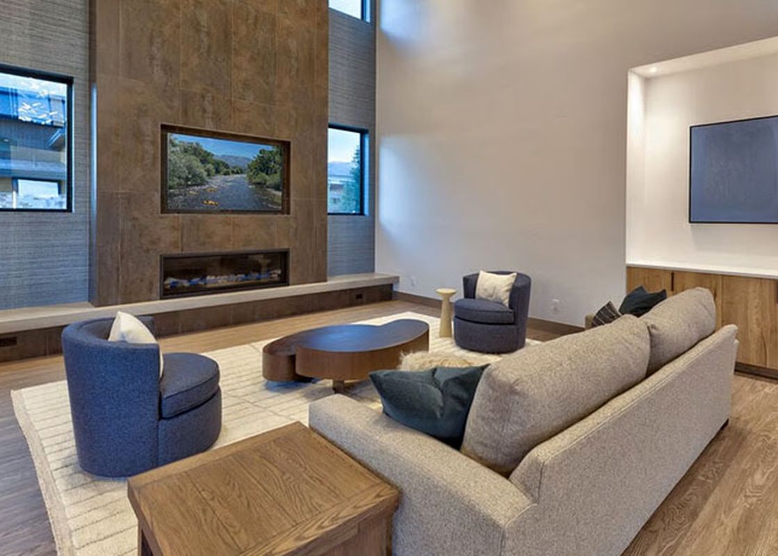 A modern and luxurious living room featuring a large gray sectional sofa, blue armchairs, a distinctive wooden coffee table, a sleek fireplace, and expansive windows that offer captivating views of the outdoors.