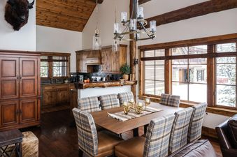 A rustic dining area with a wooden table, plaid chairs, a grand chandelier, and an open-concept kitchen, featuring a taxidermy bison head on the wall and large windows overlooking a snowy landscape.