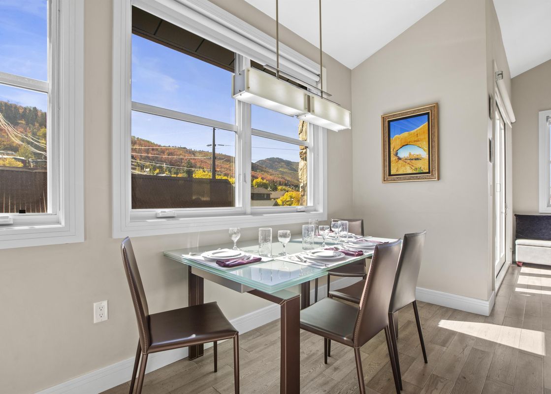 A modern dining area with a glass-top table set for four, featuring brown leather chairs, under a rectangular chandelier, with large windows overlooking autumn mountain scenery.