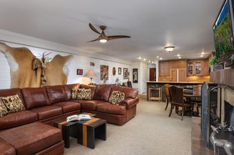 A spacious living room at Montaneros 204 features a brown leather sectional, a unique coffee table, and a well-appointed kitchen with wooden cabinetry and granite countertops, creating a cozy and inviting atmosphere.