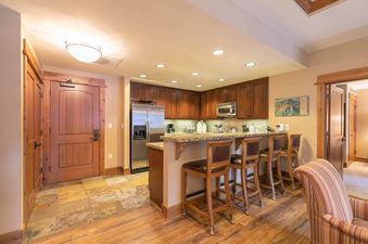 A kitchen with wood cabinets, stainless steel appliances, and a granite countertop with a bar and stools. There is a large wooden door to the right of the image and another wooden door that leads to another room.