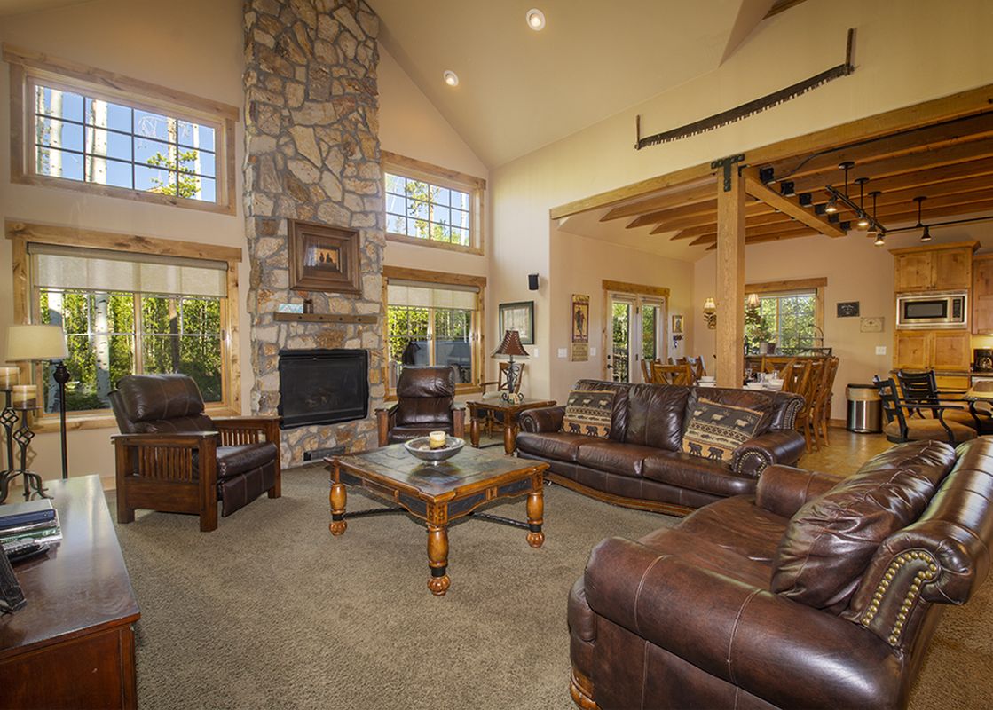 A spacious and inviting living room at Leland Creek Home, featuring comfortable leather sofas and armchairs, a large stone fireplace, and abundant natural light from numerous windows overlooking a lush, wooded exterior.