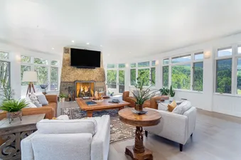A spacious and bright living room at Turkey Creek features a large stone fireplace, comfortable seating, and numerous windows offering views of a lush, green landscape.