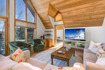 A cozy, sunlit living room at Golden Spruce Jackson Hole with vaulted wood ceilings, a large TV, comfortable sofas, and forest green armchairs.