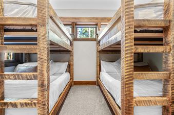 A cozy bunk room at Timberline Lodge features two sets of rustic wooden bunk beds with white bedding, separated by a central carpeted aisle leading to a window with an exterior view.