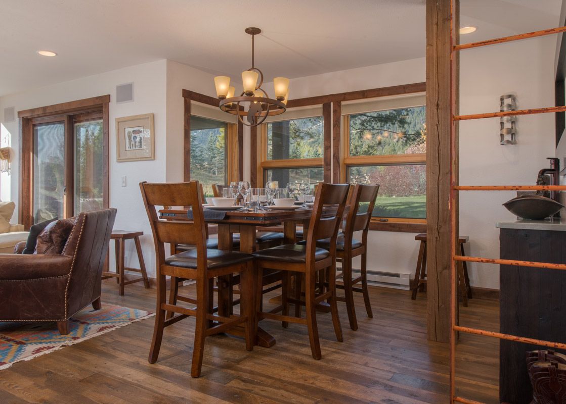 The dining area of Nez Perce D2 with a wooden table set for six, surrounded by dark chairs and illuminated by a chandelier, with large windows overlooking a scenic outdoor landscape.