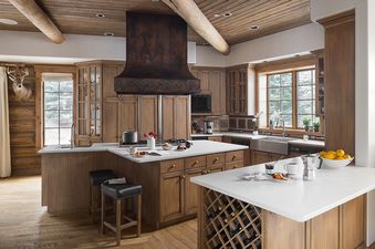 A rustic-modern kitchen featuring custom wooden cabinetry, white countertops, and a prominent dark metal range hood, with a large window overlooking a winter landscape.