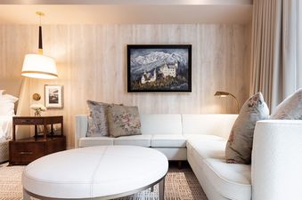 A white sectional sofa with pillows sits in a room with a wooden wall and a painting of mountains and a castle.