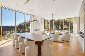 A wide shot of the dining and living area in Founders Place 1704, featuring a long wooden dining table with upholstered chairs, and a living area with curved white sofas, all set against a backdrop of large windows offering mountain views.