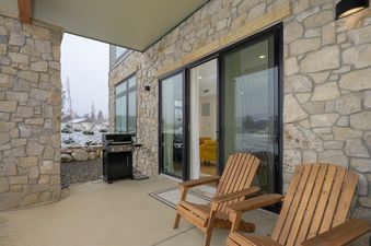 The outdoor patio features a stone-clad wall, two wooden Adirondack chairs, a black BBQ grill, and a sliding glass door leading indoors, with a snowy landscape visible in the background.