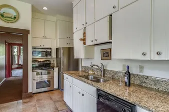 The kitchen at Summer Haus features white cabinetry, granite countertops, stainless steel appliances, and a double-basin sink, with a glimpse into an adjacent room through an open doorway.