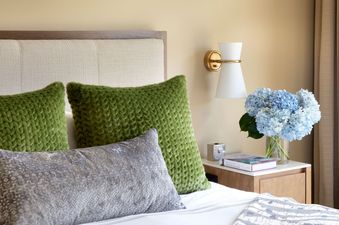 A bedroom with green pillows on a king-size bed with a white bedspread and a nightstand with a book, a clock, and a vase of flowers.