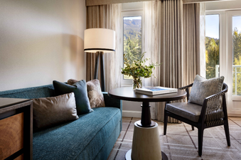A cozy corner of the Fairmont Chateau Queen and Queen Room, featuring a comfortable teal sofa, a dark wooden table with a floral arrangement, and a modern floor lamp, all set against a backdrop of a large window offering a scenic view of lush mountains.