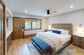 A bedroom with a large bed and a bench at the foot of the bed. The bedroom has a window with a view of the forest, a wooden dresser, and a lamp on a nightstand.
