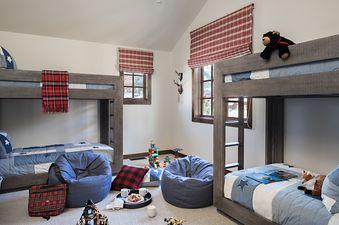 A cozy kids' bunk room at Four Pines 05 featuring two rustic wooden bunk beds with blue and white star-patterned quilts, red and white plaid window shades, two blue beanbag chairs, and colorful toys on a light-colored carpet.